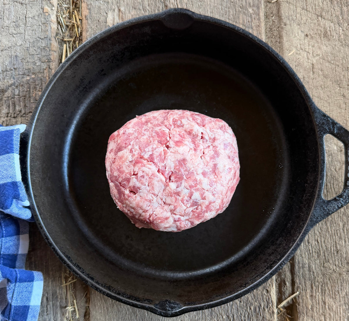 1 lb of Low PUFA corn and soy free ground pork sits inside a black cast iron skillet on a rustic wooden surface, with a blue and white checkered cloth partially visible on the left.