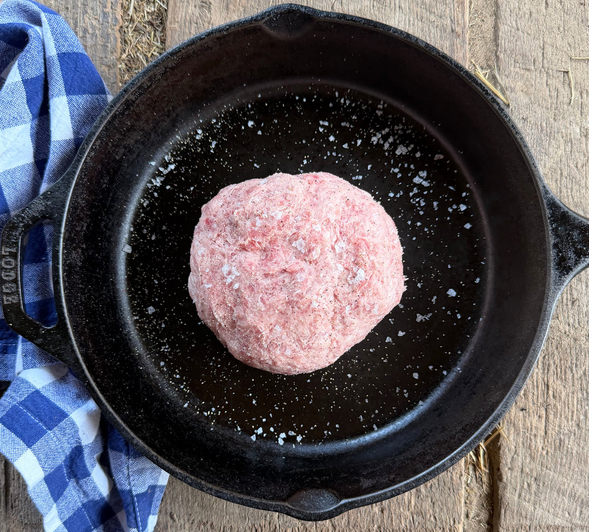 Corn and soy free raw ground salt and pepper pork sits in a black cast iron skillet on a rustic wooden table, with a blue and white checkered cloth beside the pan.