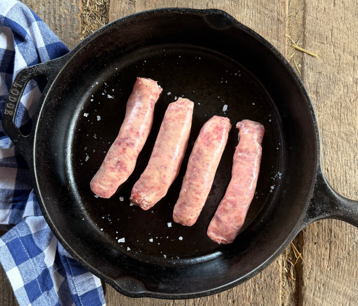 Four Low PUFA corn and soy free pork sausage links cooking in a black cast iron skillet on a rustic wooden table, with a blue and white checkered cloth beside the pan.